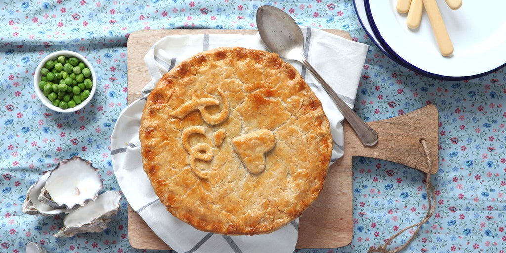 Steak, oyster and Guinness pie on a table with oyster shells and a pot of peas 