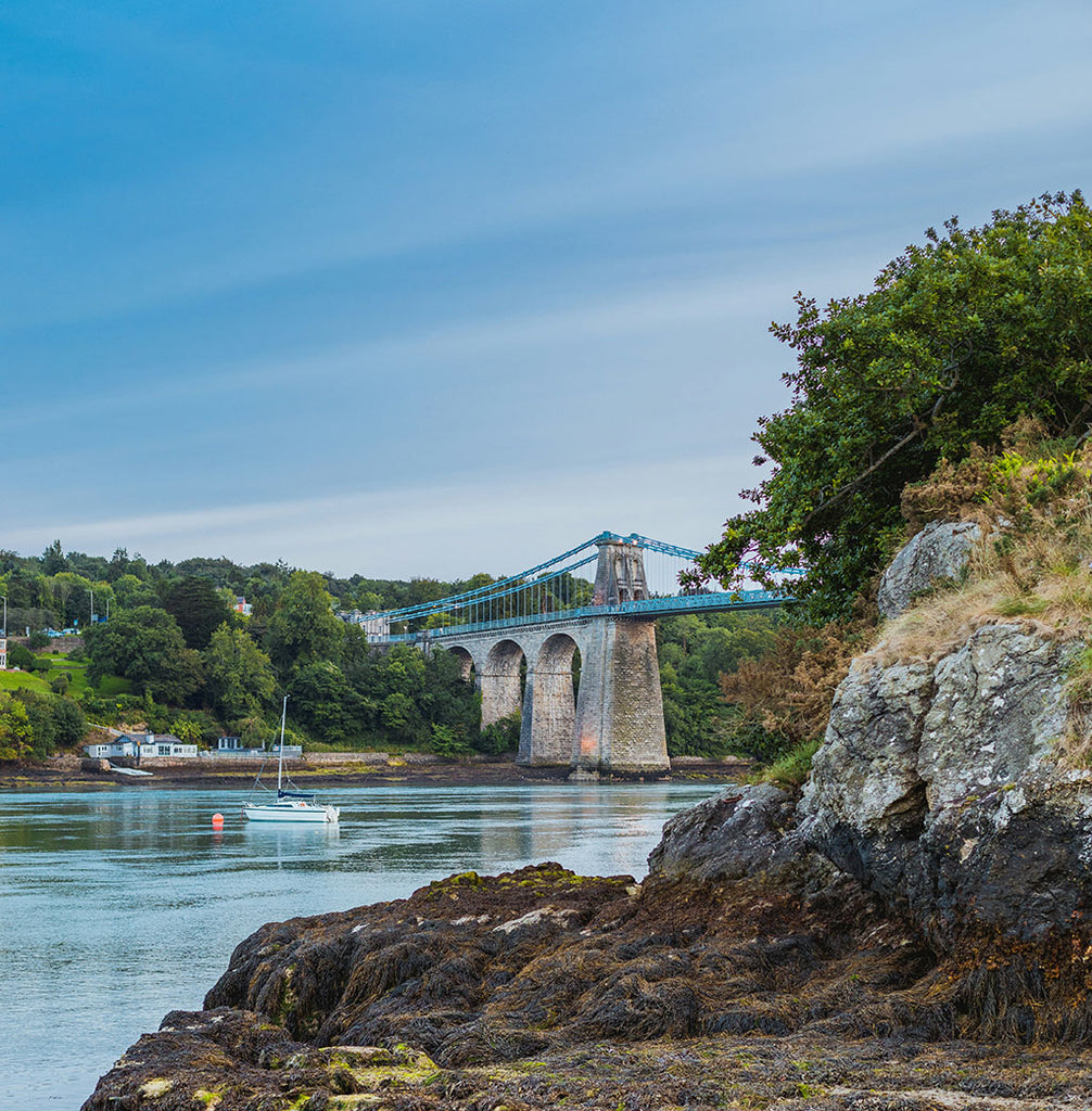 Bridge over a calm body of water with greenery and rocks in the foreground